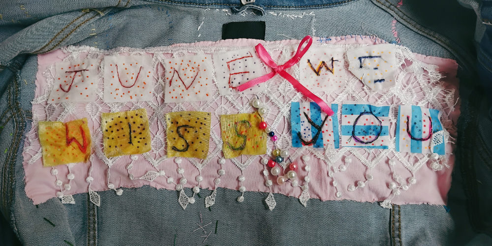 A close up of the patch - a pink rectangle of fabric with the letters We Miss You June on white, yellow and blue fabric squares