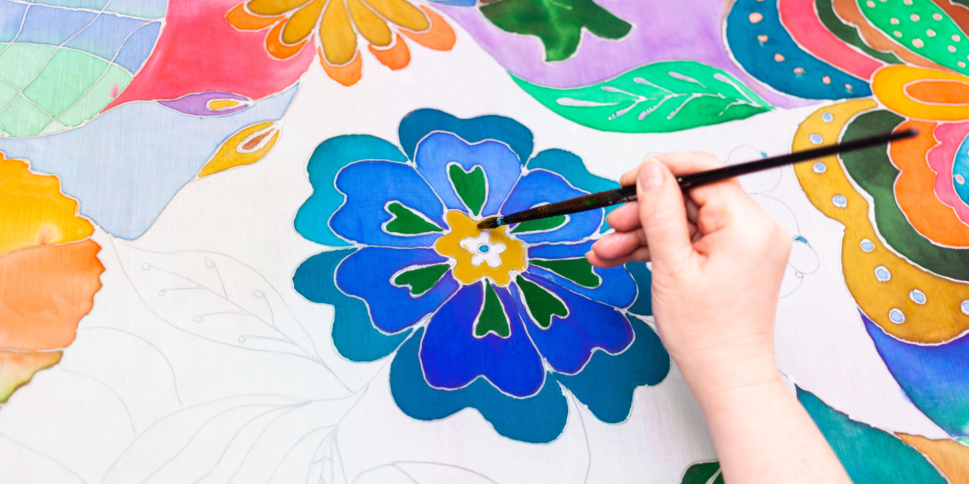 A white person's hand holds a paintbrush over a painted flower coloured in blue and yellow