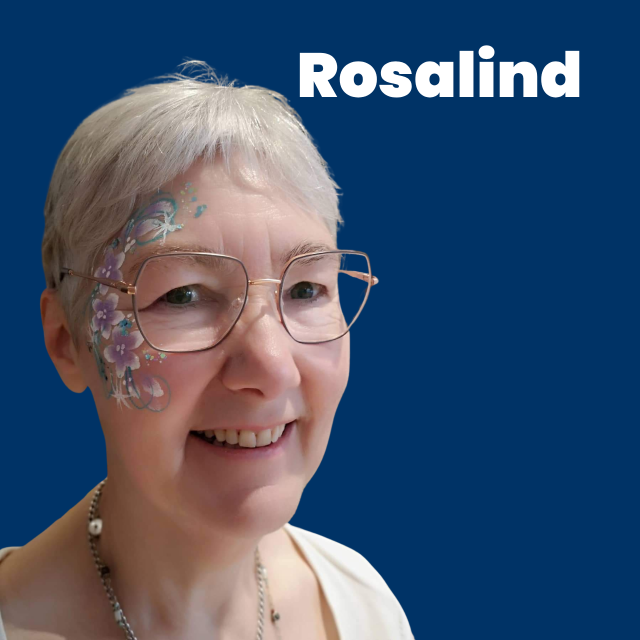 A white woman with short cropped grey hair wearing metal frame glasses and smiling. They have lilac and white flower facepaint on one side of their face. The name Rosalind on a dark blue background